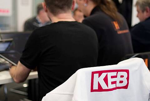 Menschen, die um einen Tisch sitzen und an Laptops arbeiten, im Vordergrund ist das Logo von KEB Automation auf einer weißen Jacke zu sehen