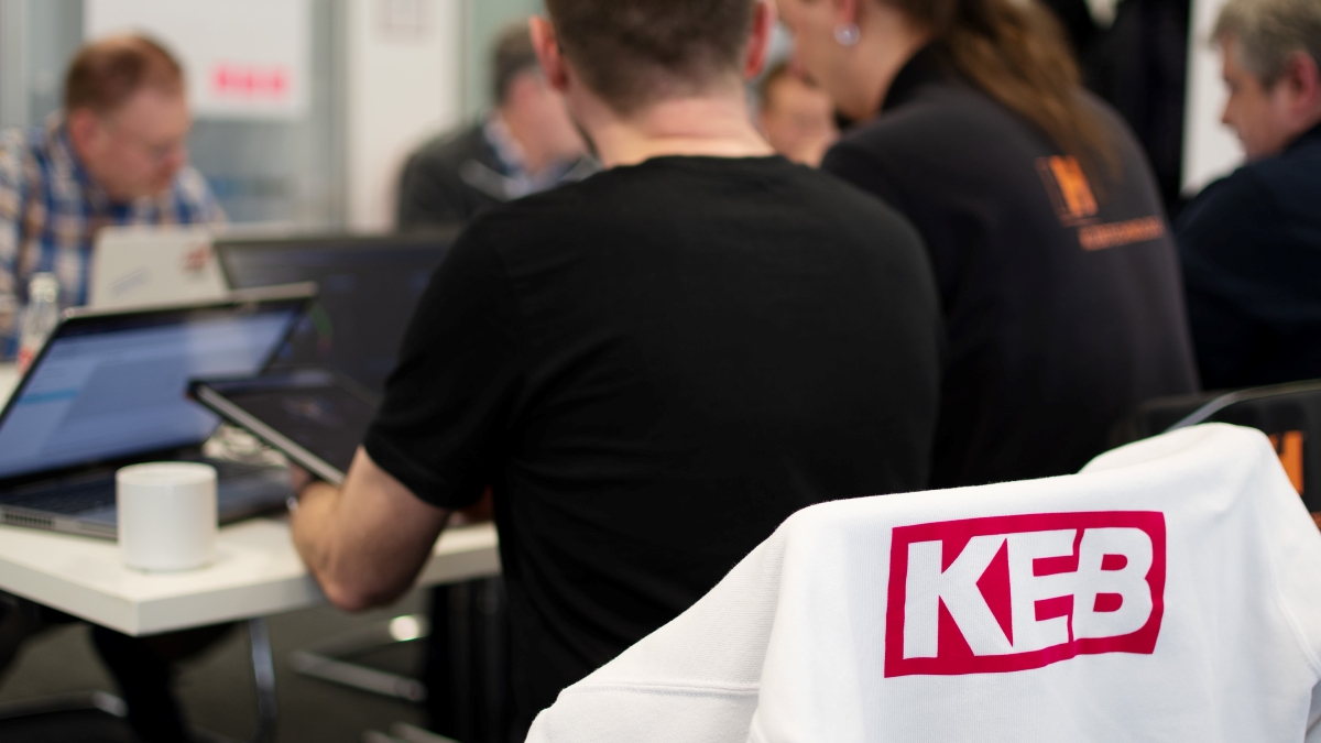 Menschen, die um einen Tisch sitzen und an Laptops arbeiten, im Vordergrund ist das Logo von KEB Automation auf einer weißen Jacke zu sehen